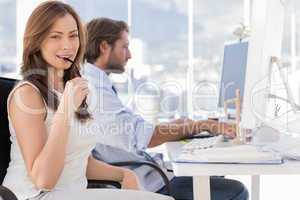Woman biting her glasses with colleague working behind