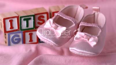 Baby booties falling on pink blanket with its a girl message in blocks