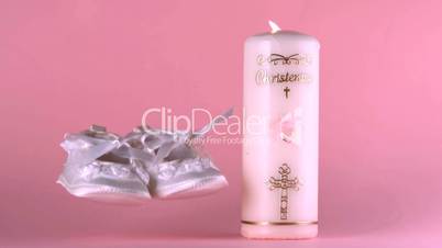 Baby shoes falling beside lit baptism candle on pink background