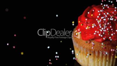 Colourful sprinkles pouring onto cupcake on black surface