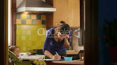 Multi-tasking mom cooking and working, feeding child in home kitchen