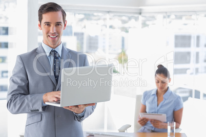 Businessman using laptop standing in office smiling at camera
