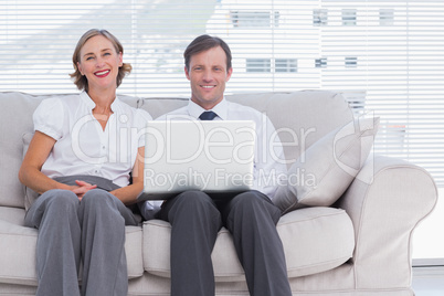 Two colleagues sitting on couch using laptop in bright office