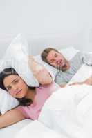 Upset woman covering ears with pillow next to husband snoring