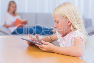 Little girl using tablet while her mother is reading on the couc