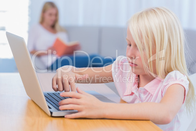Little girl using laptop while her mother is reading on the couc