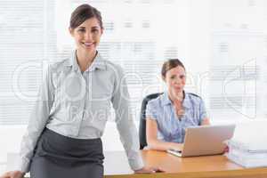 Businesswomen smiling at camera with one standing and one sittin