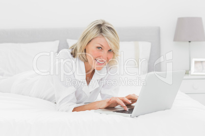 Smiling woman using her laptop on her bed looking at camera
