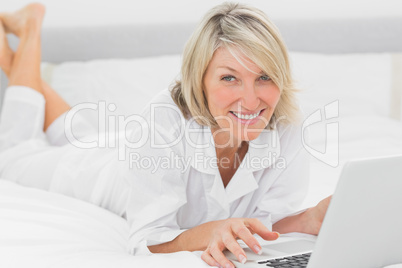 Happy woman using her laptop on her bed smiling at camera