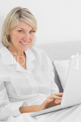 Cheerful woman sitting in bed with laptop looking at camera