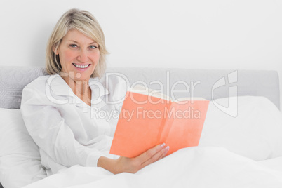 Blonde woman sitting in bed reading smiling at camera