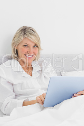 Cheerful woman sitting in bed using tablet pc looking at camera