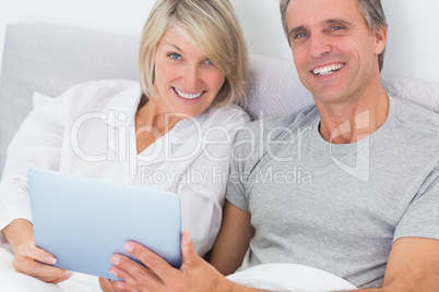 Couple using tablet pc in bed smiling at camera