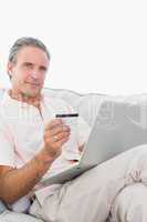 Man on his couch using laptop for shopping online smiling at cam