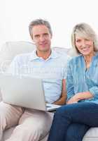 Couple using laptop together on the couch smiling at camera