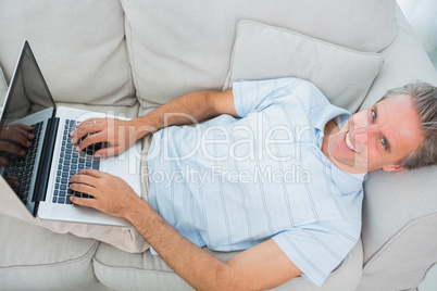Man lying on couch typing on laptop smiling at camera