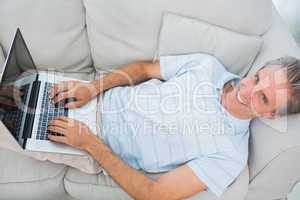 Man lying on couch typing on laptop smiling at camera