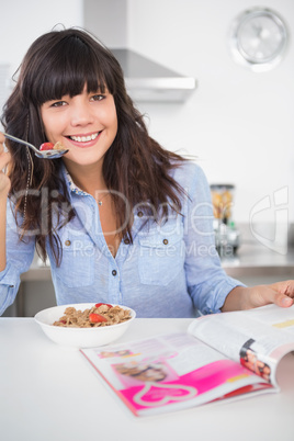 Cheerful brunette eating cereal and reading magazine