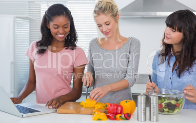Cheerful friends making salad and using laptop for recipe