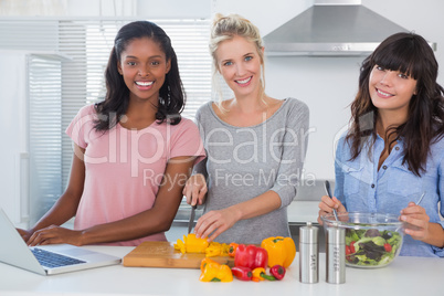 Cheerful friends making salad and using laptop for recipe lookin