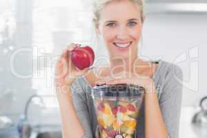 Blonde woman leaning on her juicer full of fruit and holding red