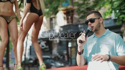 man speaks into a microphone in the background dancing girls in swimsuits