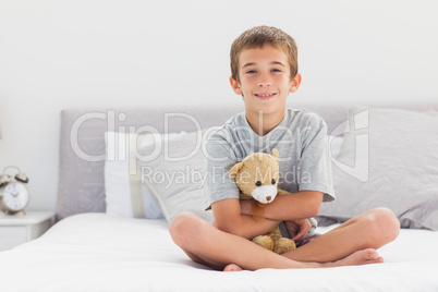 Smiling little boy sitting on bed holding his teddy bear