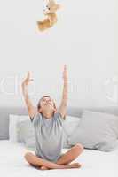 Little boy sitting on bed throwing his teddy bear to the ceiling