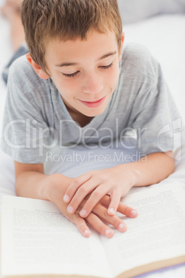 Smiling little boy lying on bed reading book