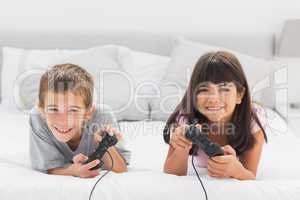 Smiling siblings lying on bed playing video games together