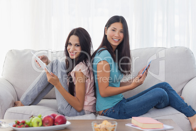 Friends sitting back to back using their tablets smiling at came