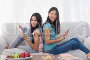 Friends sitting back to back using their tablets smiling at came