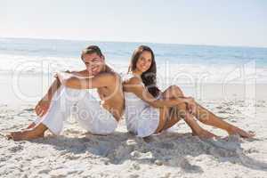 Couple sitting back to back on the sand smiling at camera