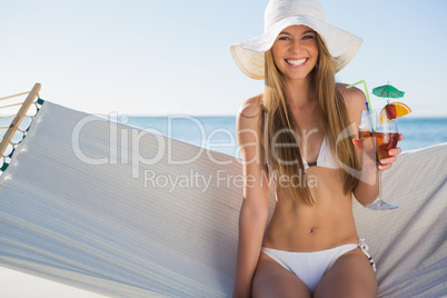 Happy blonde wearing bikini and sunhat sitting on hammock with c