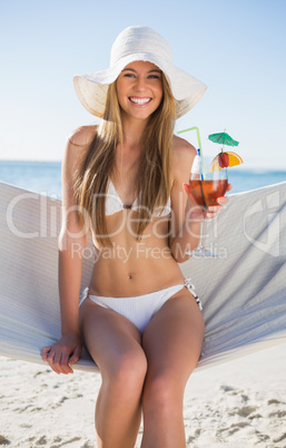 Smiling blonde wearing bikini and sunhat sitting on hammock with