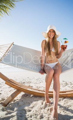 Cheerful blonde wearing bikini and sunhat sitting on hammock wit
