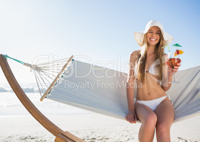 Smiling blonde wearing sunhat sitting on hammock with cocktail