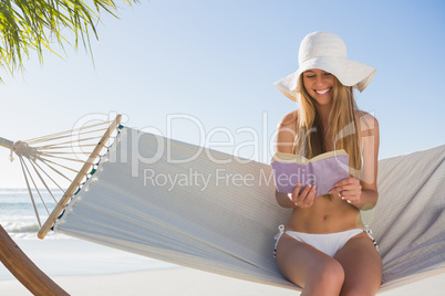 Smiling blonde wearing sunhat sitting on hammock reading book