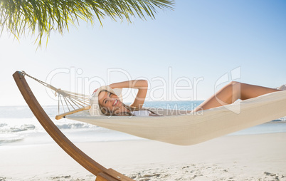 Woman wearing sunhat and bikini relaxing on hammock smiling at c