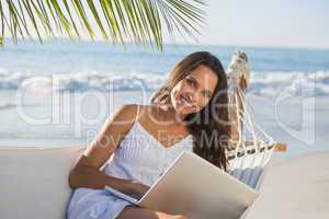 Pretty brunette sitting on hammock with laptop smiling at camera