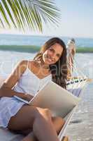 Brunette sitting on hammock using laptop smiling at camera