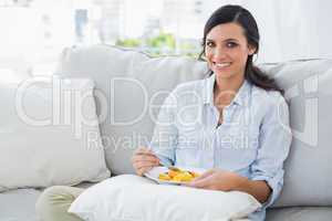 Cheerful woman sitting on the sofa eating fruit salad