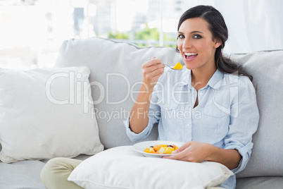 Happy woman sitting on the sofa eating fruit salad