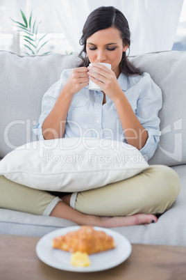Cute woman on sofa drinking coffee and having croissant