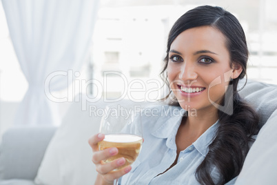 Happy pretty brunette drinking white wine sitting on sofa