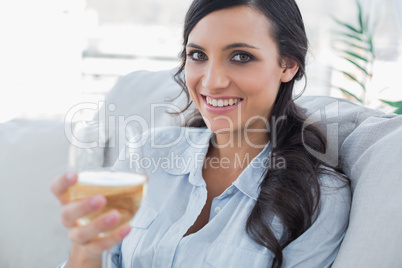 Cheerful pretty brunette drinking white wine sitting on sofa