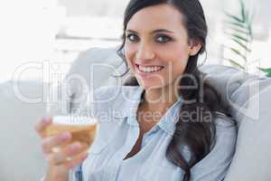 Cheerful pretty brunette drinking white wine sitting on sofa