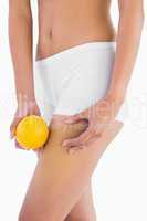 Slender woman squeezing fat on thigh as she holds orange