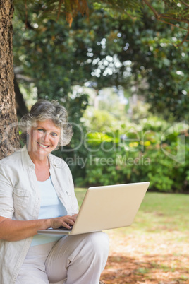 Happy mature woman using a laptop sitting on tree trunk Happy mature woman using a laptop sitting on tree trunk