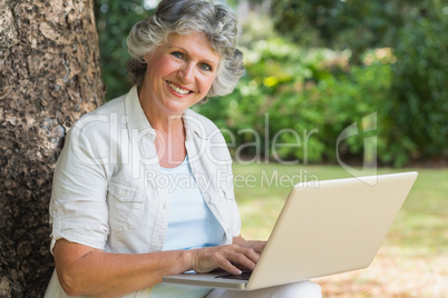 Cheerful mature woman using a laptop sitting on tree trunk